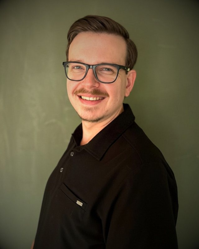 Alex Adamczyk, LMT, smiling, in a black work shirt