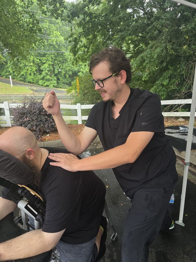 Alex Adamczyk, LMT, working on a client's shoulders in a massage chair outdoors at Hustle House