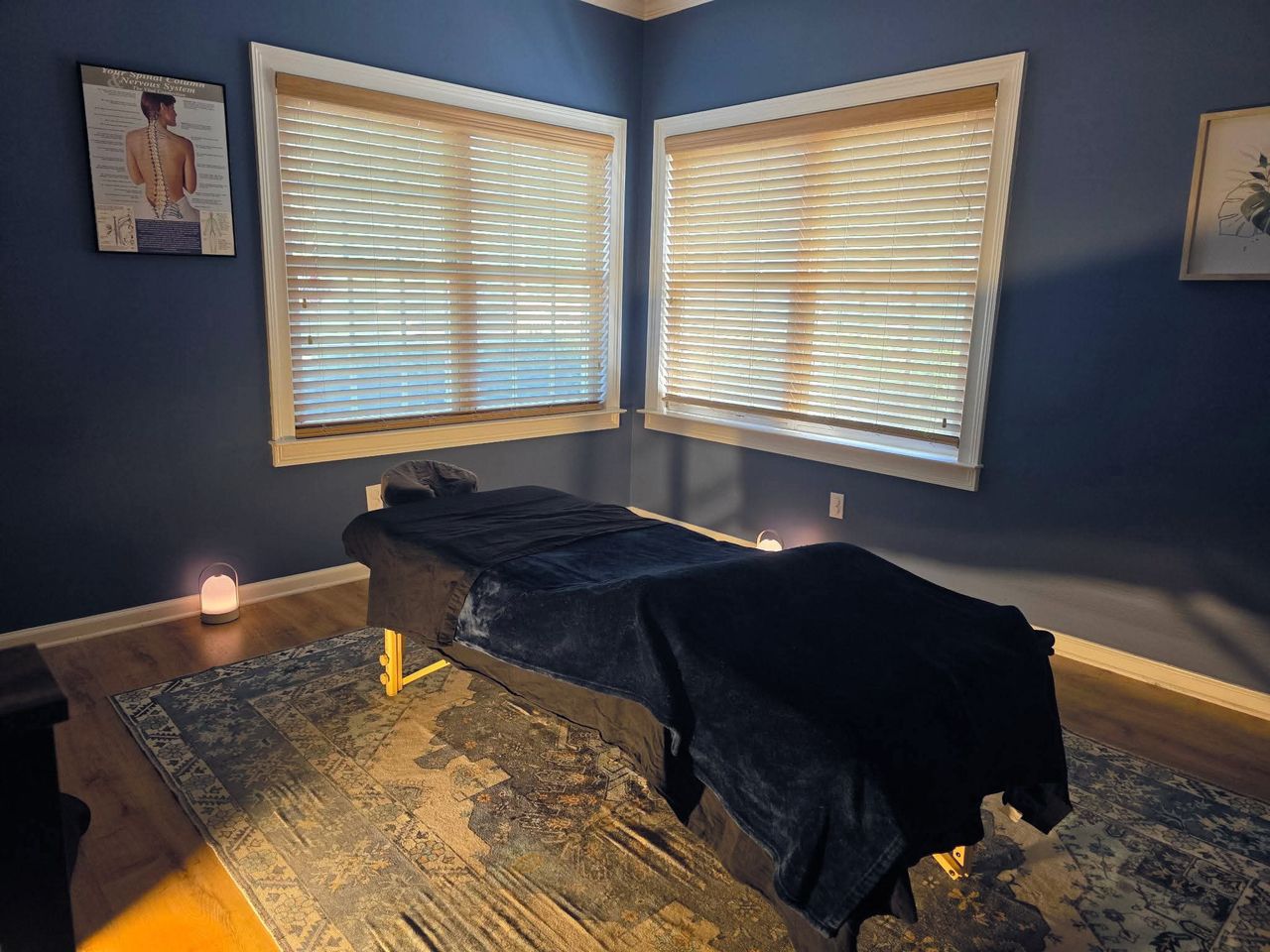 The whole Elbow Room treatment room: a massage table with a navy blanket under two windows with wood blinds, a lantern glowing on the floor, and a spine chart on the wall