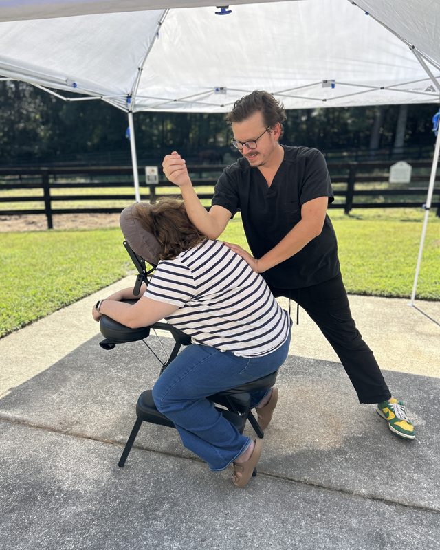 Alex Adamczyk, LMT, working on a seated person's shoulder with his forearm during a chair massage under a canopy at the Cherokee Center for Change, with the farm fence behind